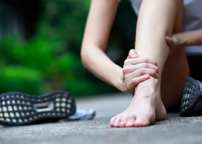 Person sitting outdoors holding their lower leg and ankle, indicating ankle pain or injury.