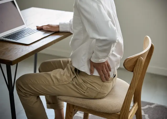 Person sitting at a desk and holding their lower back, indicating workplace‑related back pain or poor posture.
