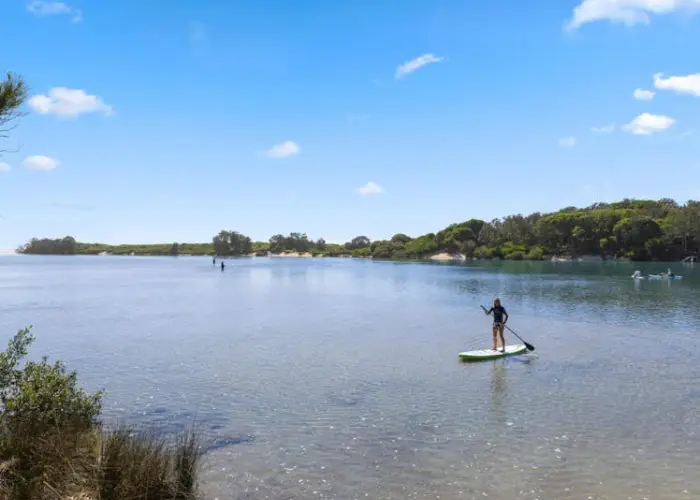 Paddleboarder on calm water in Valla surrounded by greenery and clear blue sky.